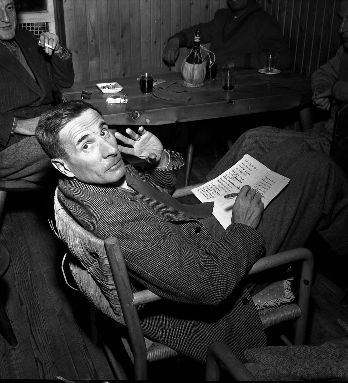 El autor, Dino Buzzati, corrige un texto en una taberna rodeado de paisanos, tabaco y chatos de vino 